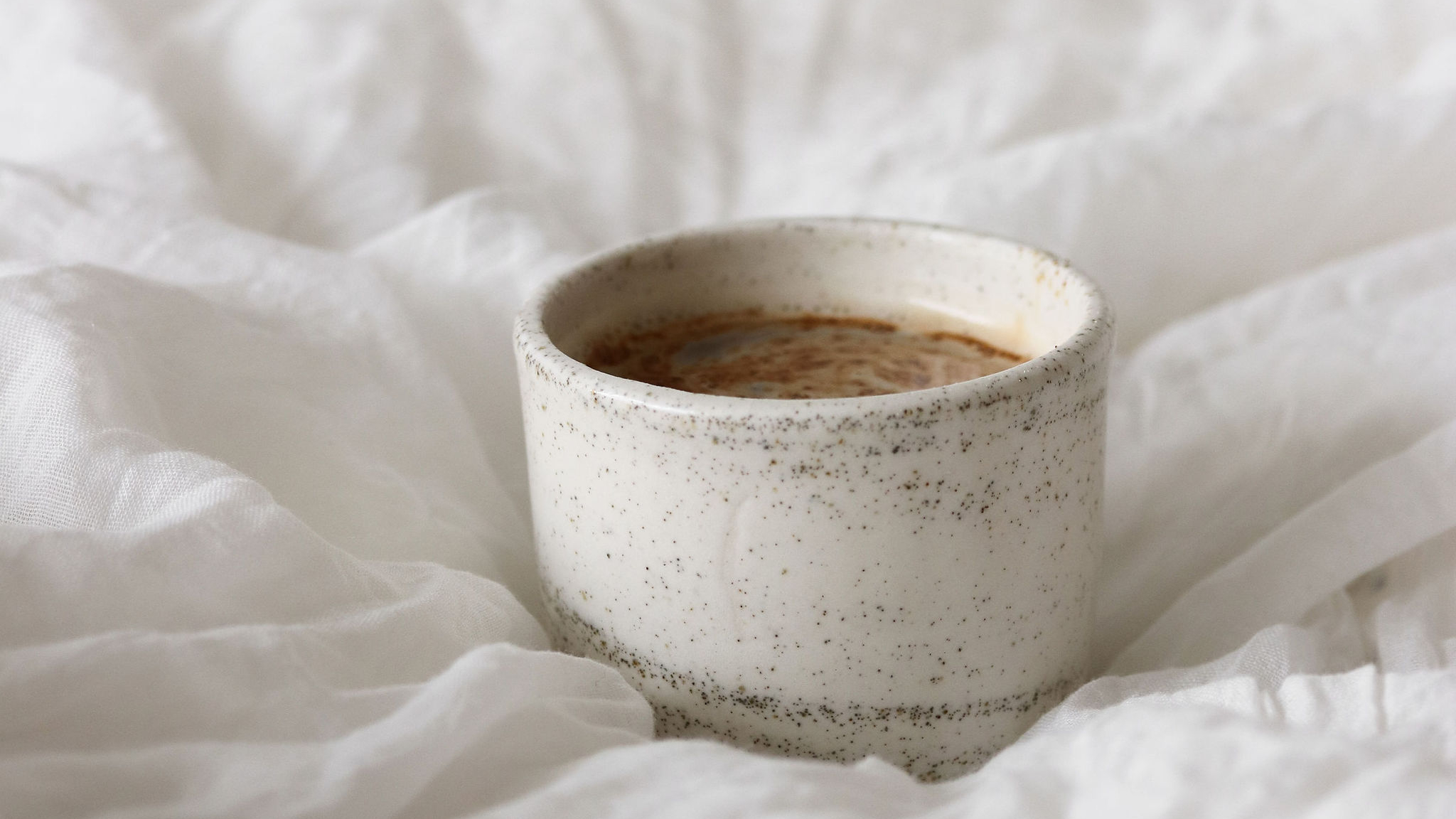Minimalist cozy moment with a warm cup of coffee in a speckled ceramic mug resting on soft white bedding. Perfect visual for a calming morning and gentle boundaries theme for highly sensitive moms.
